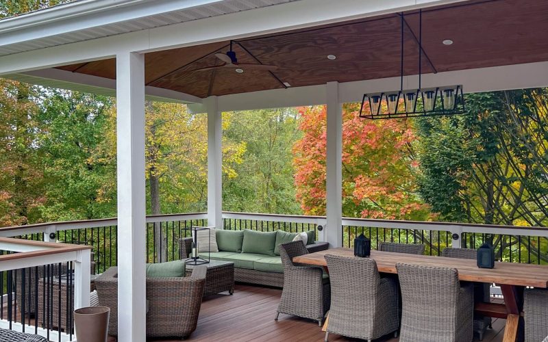A luxury elevated deck with wicker patio furniture, wood dining table with chairs, ceiling fan and modern chandelier.  Pictured in autumn with fall colors.
