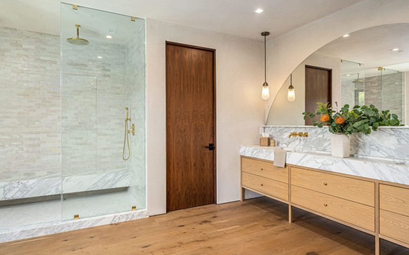 Modern bathroom interior with marble countertops, wooden cabinets, and a glass shower enclosure featuring elegant lighting and decor.